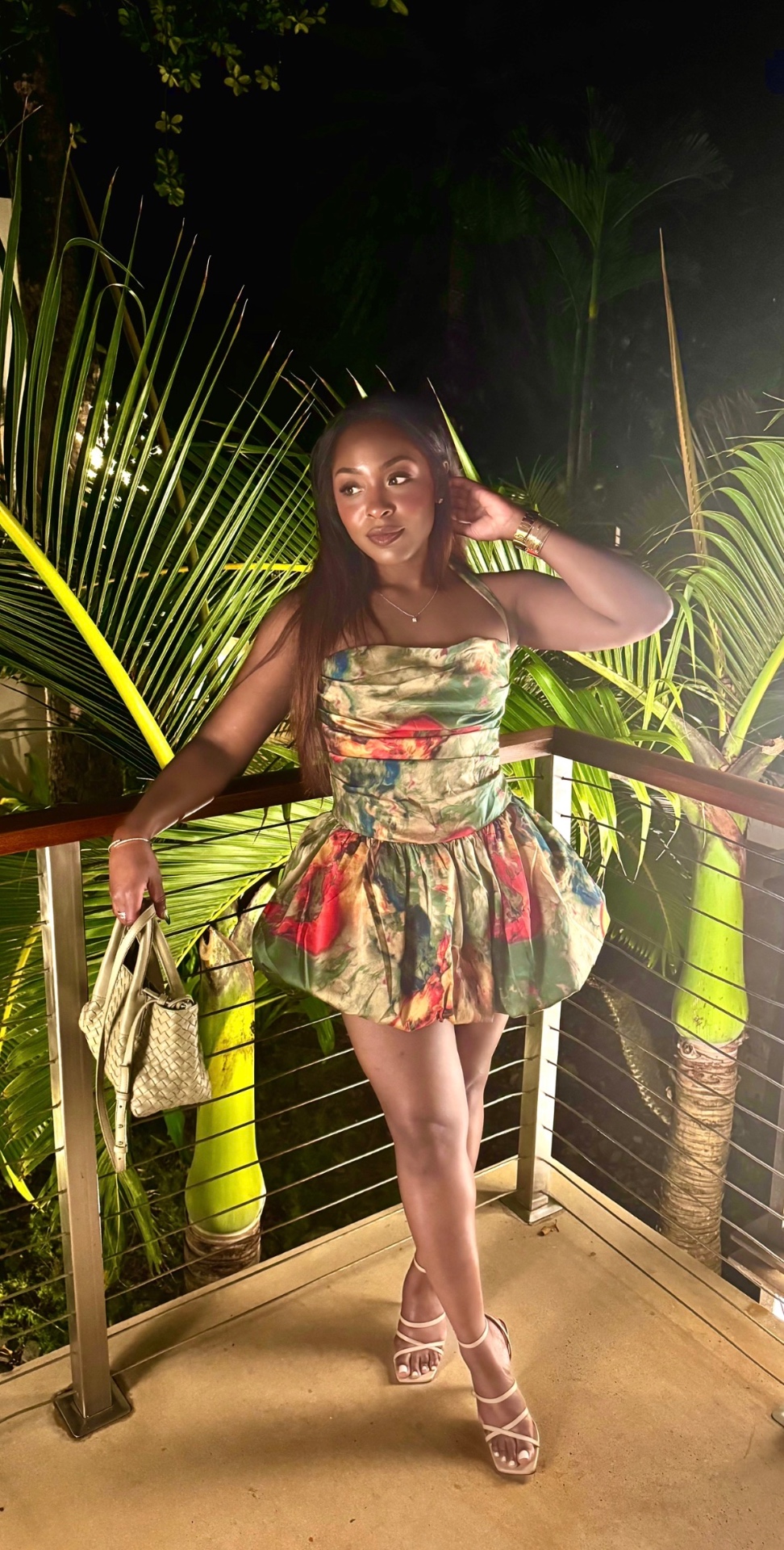 A woman in a floral strapless dress standing on a balcony beside illuminated palm leaves at night